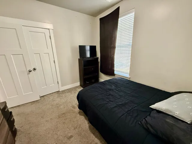 Carpeted flooring with light-toned walls, featuring a window with horizontal blinds and a built-in closet with double panel doors