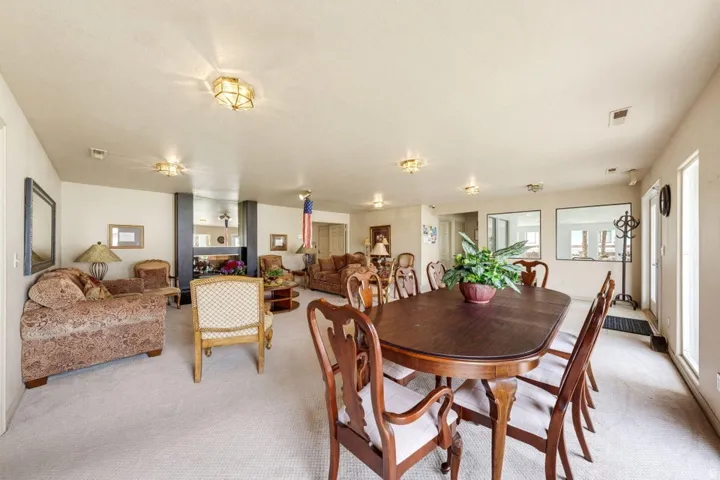 Spacious living area featuring light-colored carpeting, multiple ceiling-mounted light fixtures, a prominent fireplace with a dark finish, and two large windows