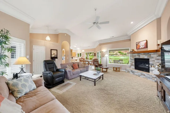 Spacious living area featuring a stone-clad fireplace with wood mantel, vaulted ceilings, crown molding, and large windows with plantation shutters