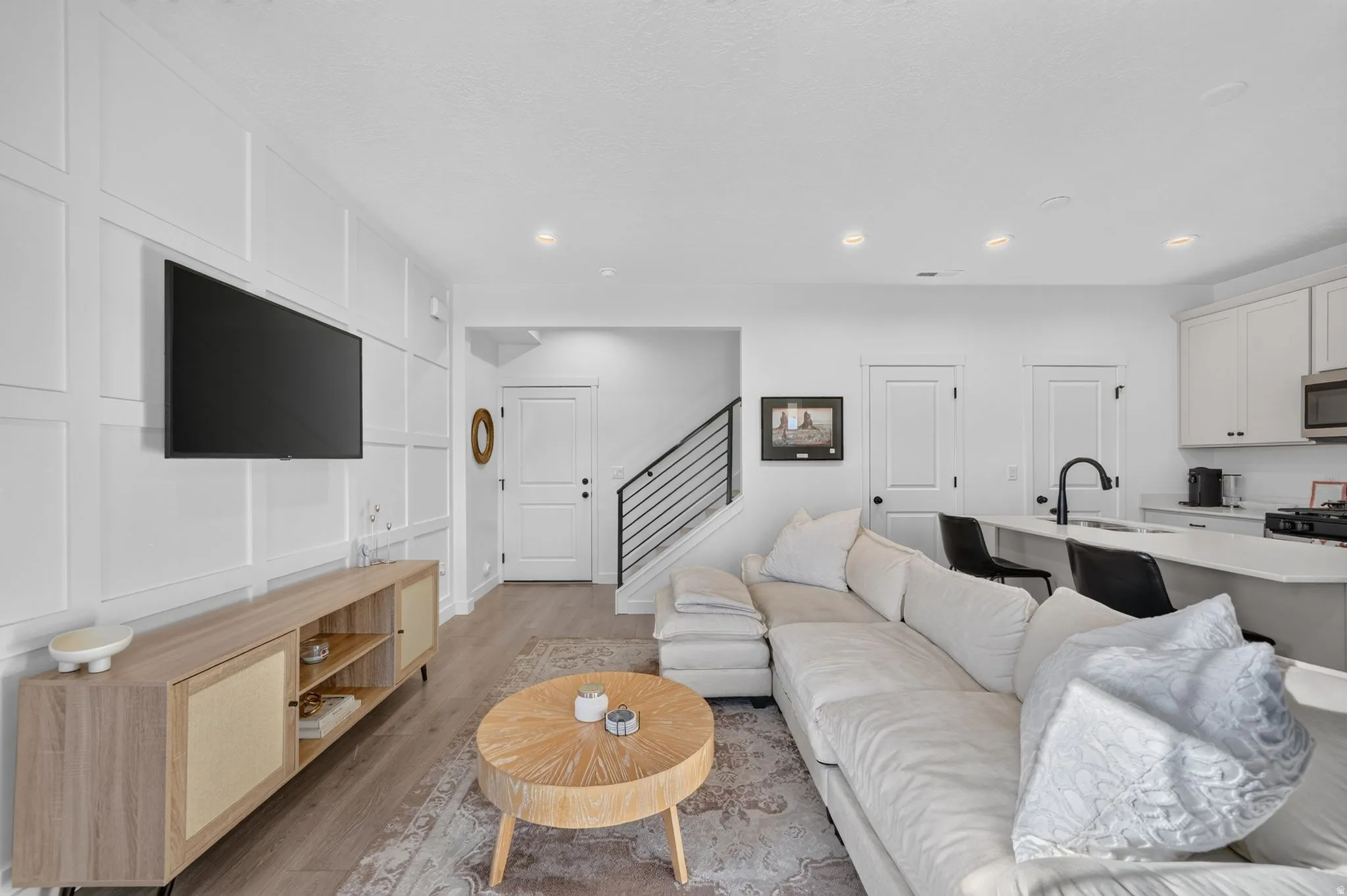 Modern living area featuring wood-finish flooring, white paneled accent wall, and a kitchen with white cabinetry