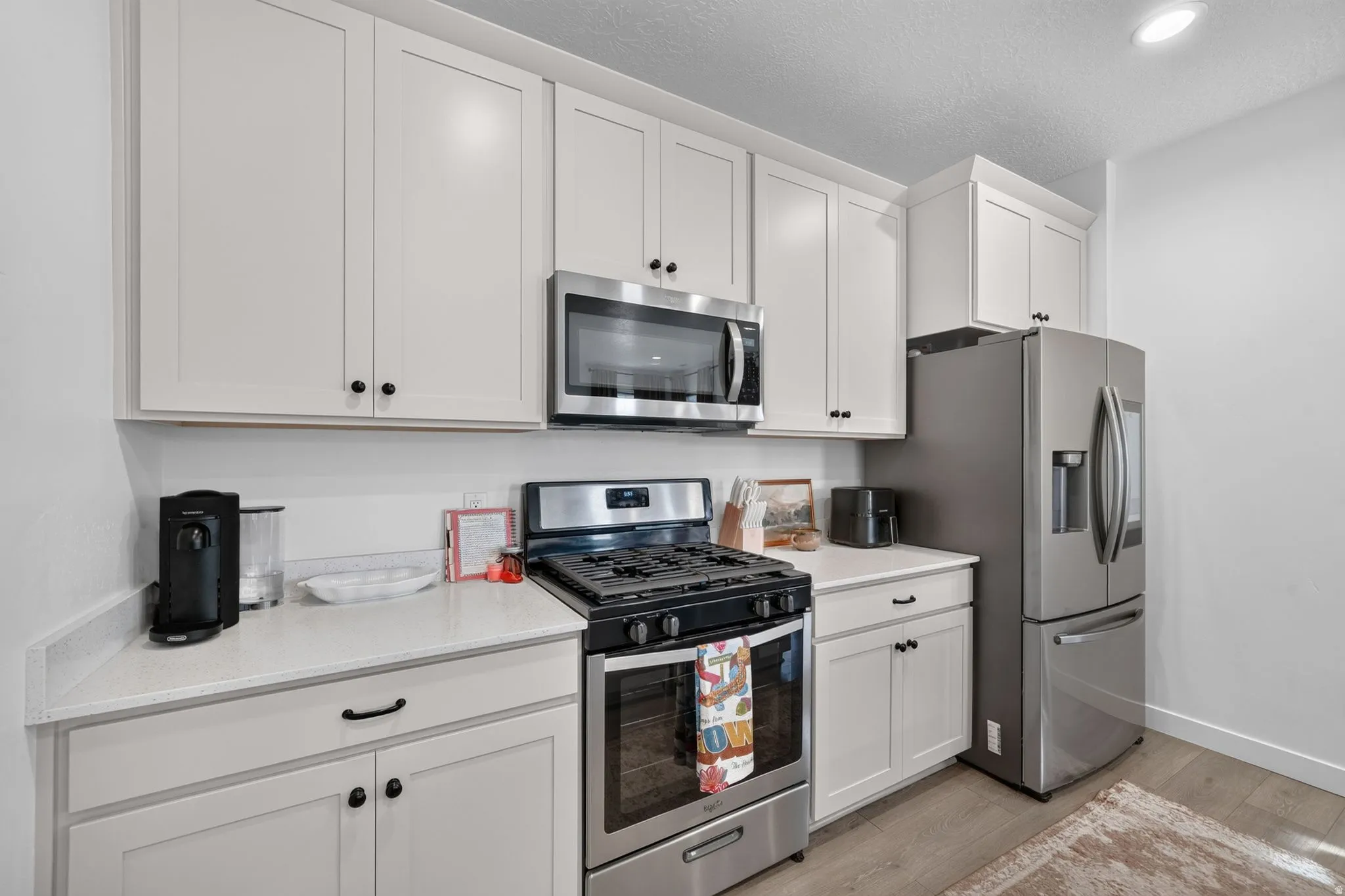 White shaker-style cabinetry, stainless steel appliances, light-toned countertops, and light wood-finish flooring
