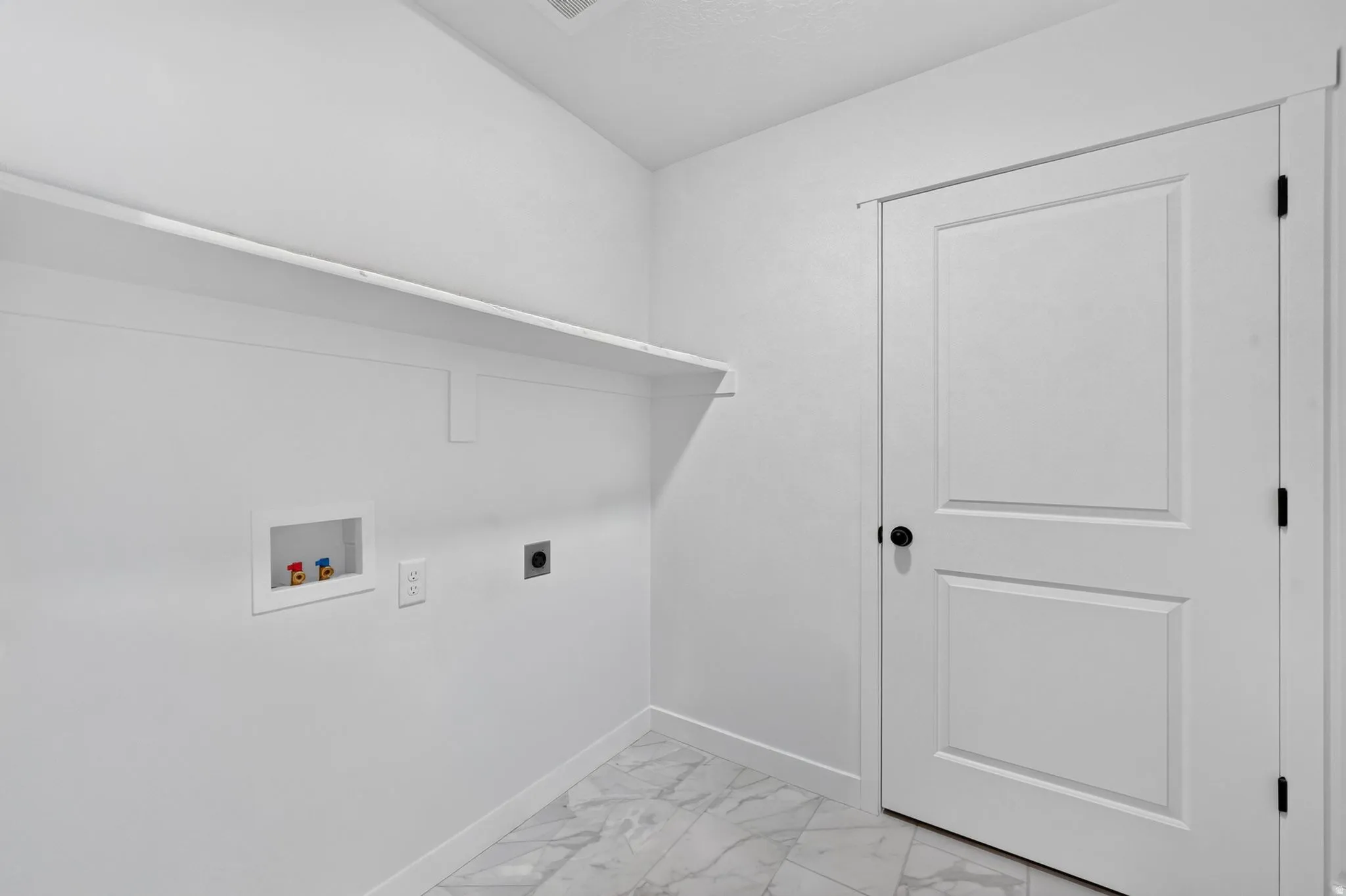 Bright laundry room featuring polished marble-finish flooring, white shaker-style cabinetry with integrated shelving, recessed electrical outlets, and a two-panel white interior door with black hardware