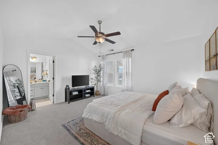 Vaulted ceiling with a ceiling fan, carpet flooring, multiple windows with blinds, and a door leading to a bathroom with a vanity