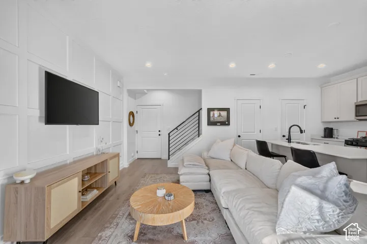 Modern living area featuring wood-finish flooring, white paneled accent wall, and a kitchen with white cabinetry
