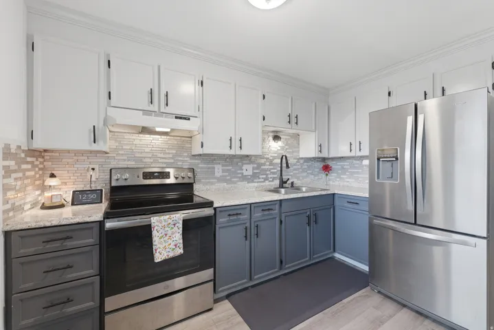 Two-tone cabinetry with white upper cabinets and gray lower cabinets, stainless steel appliances, light-toned countertop, mosaic tile backsplash, and light wood-finish flooring