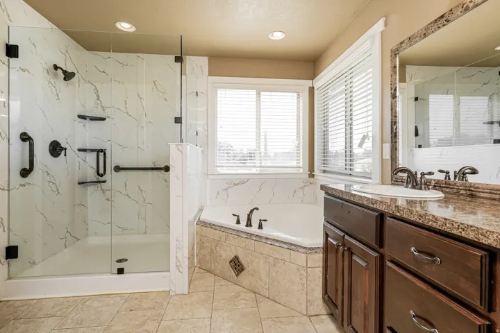 Walk-in shower with clear glass enclosure, corner soaking tub, double vanity with stone-finish countertop, and tile flooring