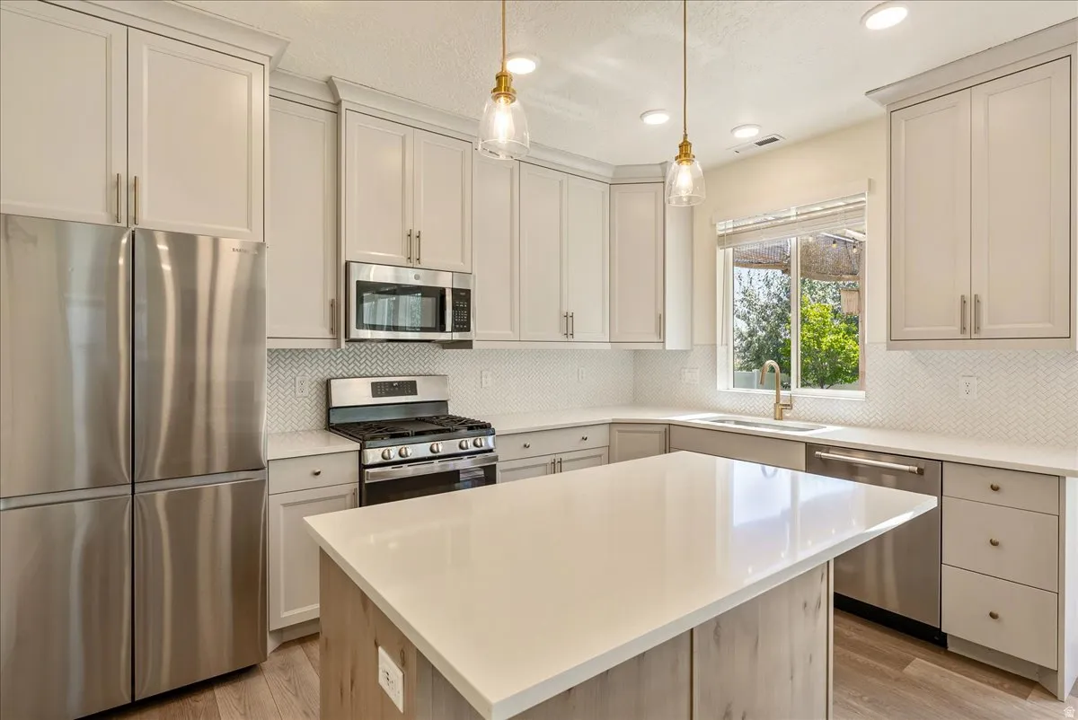 Kitchen featuring a central island with a light-toned countertop, stainless steel appliances, light-toned cabinetry, herringbone-patterned tile backsplash, and wood-finish flooring