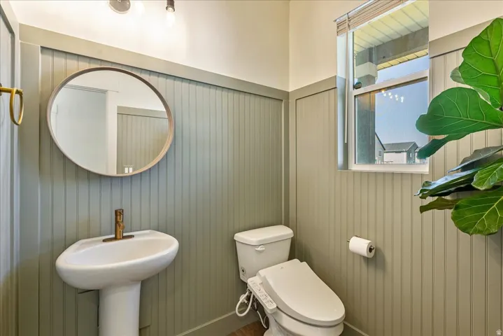Pedestal sink with brass-finish faucet, oval mirror, beadboard wall paneling, and an operable window