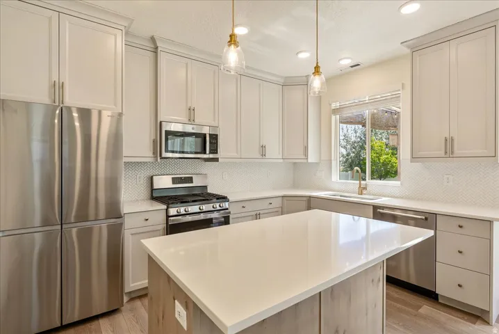 Kitchen featuring a central island with a light-toned countertop, stainless steel appliances, light-toned cabinetry, herringbone-patterned tile backsplash, and wood-finish flooring