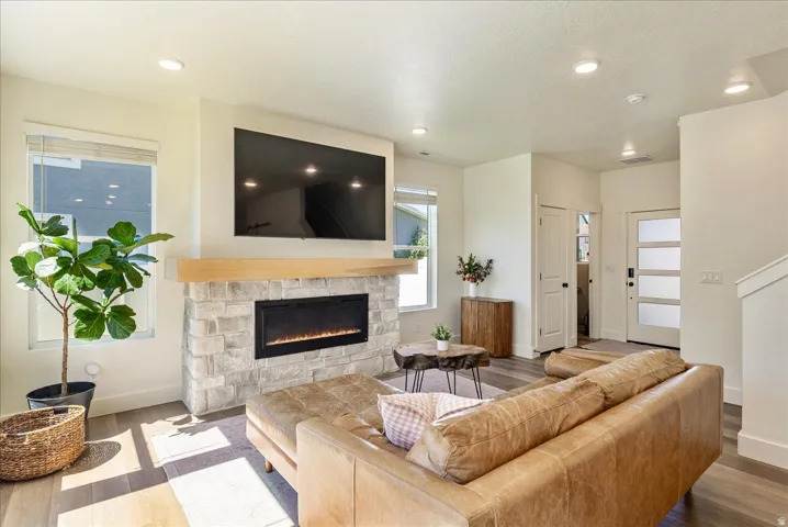 Modern living room featuring a linear electric fireplace with stone surround, wall-mounted flat-screen television, recessed ceiling lighting, hardwood flooring, and abundant natural light from multiple windows