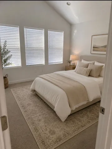 Vaulted ceiling with recessed lighting, three windows with horizontal blinds, neutral wall paint, and carpeted flooring. Virtually staged.