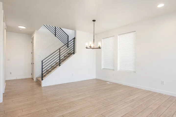 Light wood-finish flooring, staircase with metal railings, two windows with blinds, recessed lighting, and a chandelier