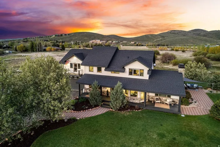 Expansive residence featuring a covered front porch, large asphalt shingle roof, stone chimney, and a paved outdoor patio