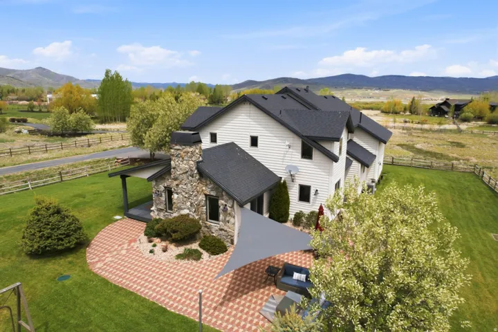 Expansive green lawn with mountain backdrop, large stone chimney, white siding exterior, and a red brick paver patio