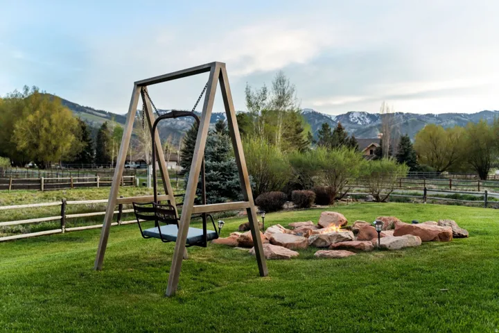 Expansive green lawn, wood-frame swing, rock-lined fire pit, mature landscaping, and split-rail fencing
