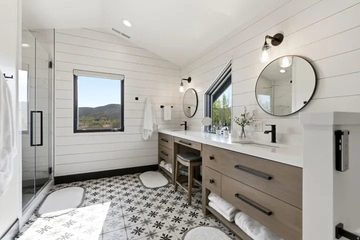 Tiled flooring with geometric patterns, dual vanity with undermount sinks, quartz countertops, black-framed windows, and a glass-enclosed shower