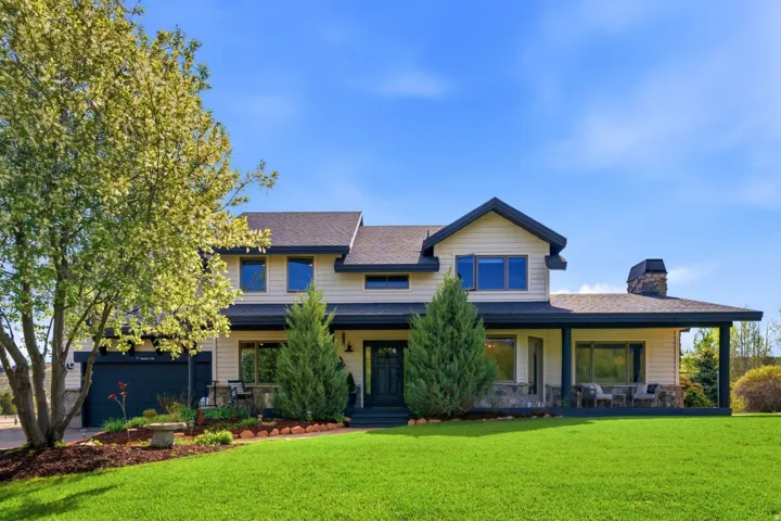 Expansive front lawn with mature landscaping, a covered front porch, stone chimney, two-car garage, and a light-colored exterior