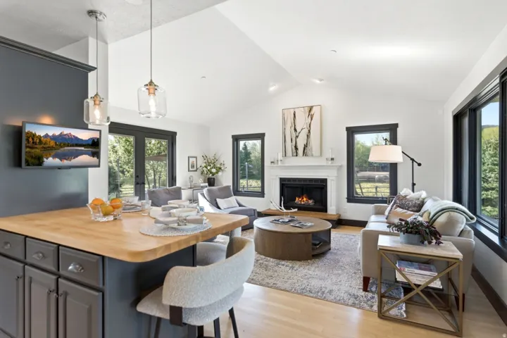 Open-concept living space with vaulted ceilings, a fireplace with a classic mantel, wood-finish flooring, and large windows with dark trim