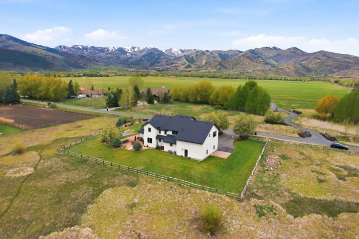 Contemporary residence with a dark roof and light-toned siding, expansive fenced yard, stone patio area, and surrounding green fields