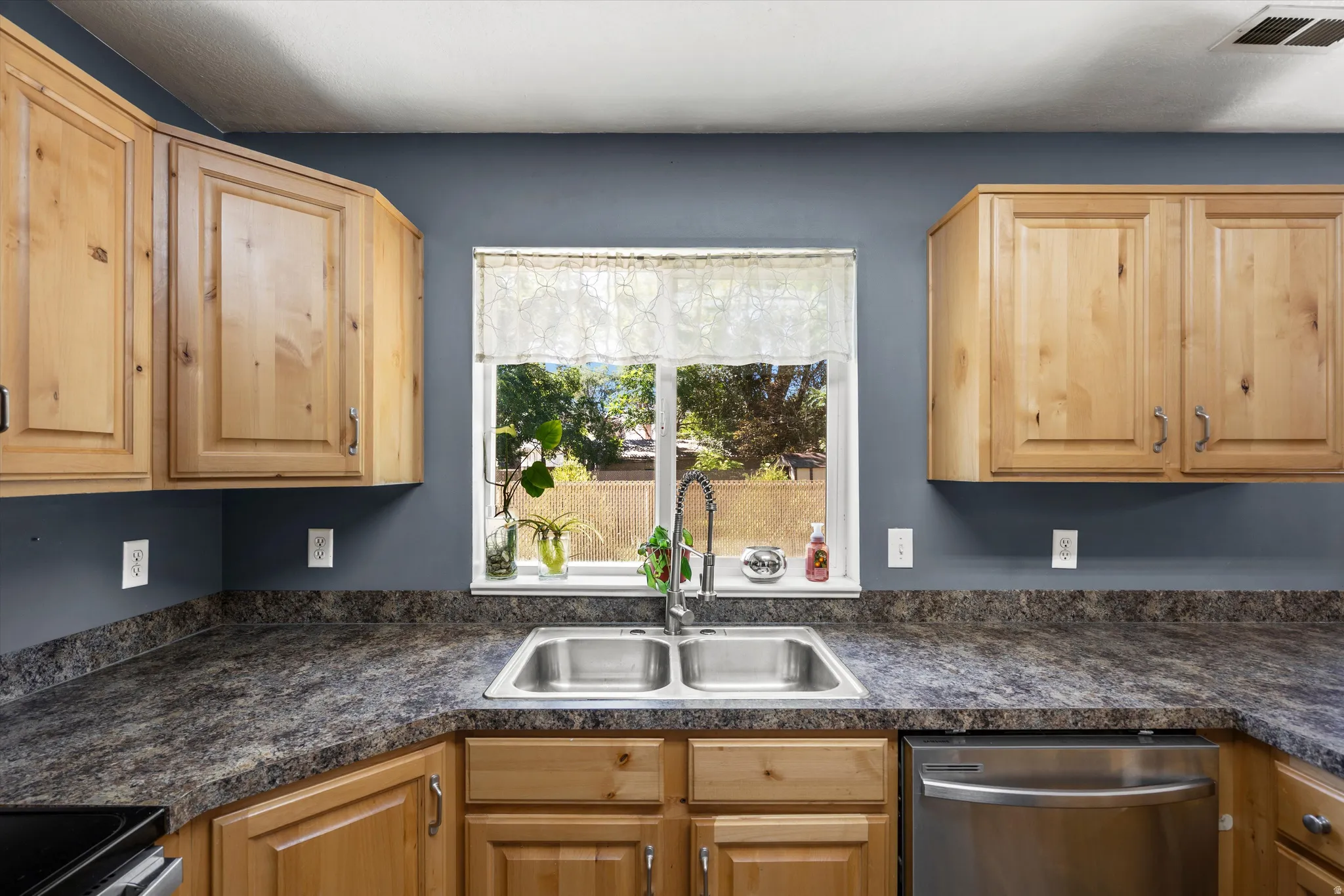 Kitchen featuring light wood cabinetry, a dual-basin stainless steel sink, a stainless steel dishwasher, and dark patterned countertops