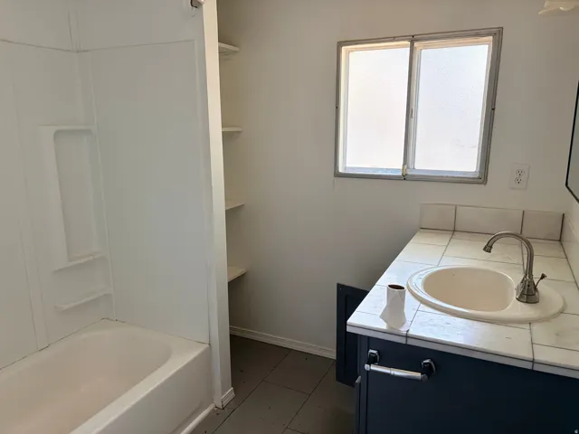 Integrated bathtub, tile countertop with a drop-in sink, dark wood-finish vanity, and built-in shelving