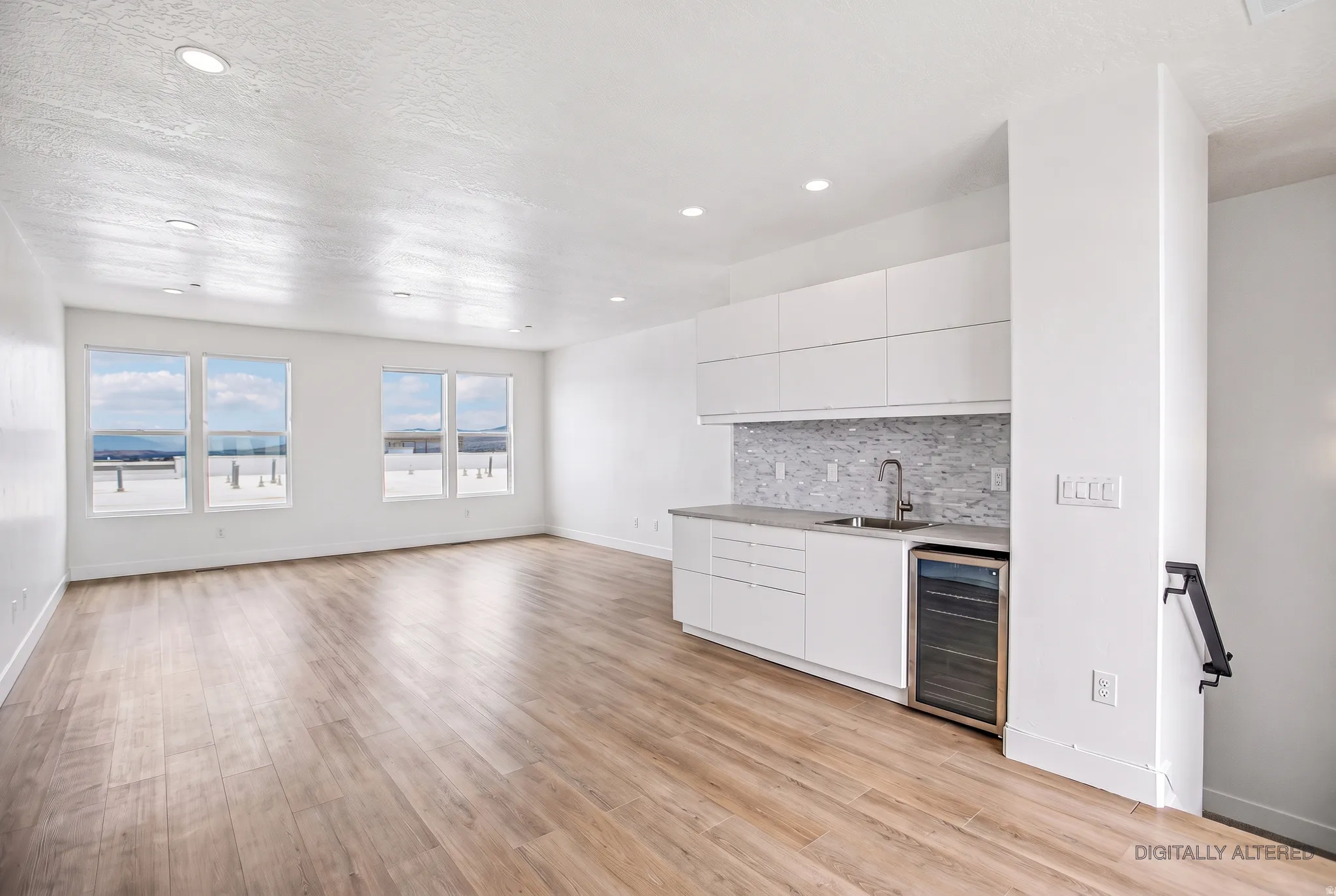 Open-concept living and kitchen space featuring gleaming wood-finish flooring throughout, expansive windows with natural light, white cabinetry with integrated appliances, gray tile backsplash, stainless steel sink and faucet, and recessed ceiling lightin