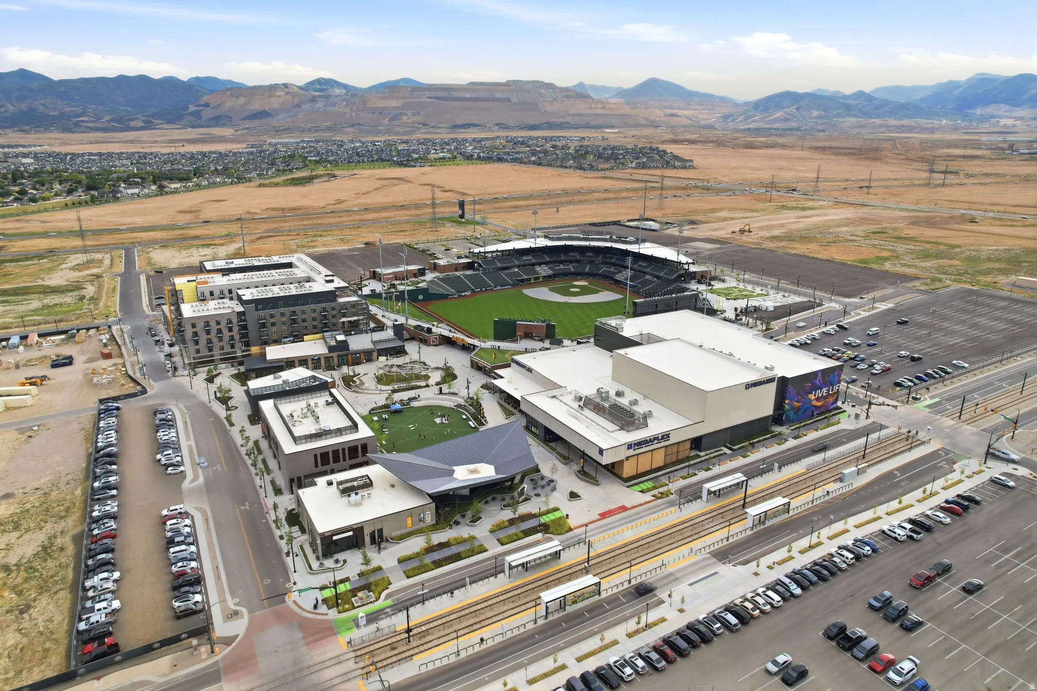 Mixed-use development featuring multi-story residential buildings, commercial structures, a baseball stadium, and a light rail system with platforms