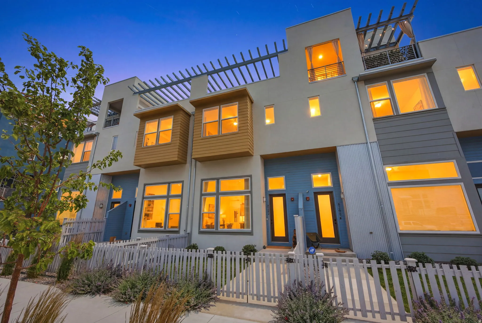 Contemporary townhomes featuring a multi-story facade, prominent windows, and a white picket fence