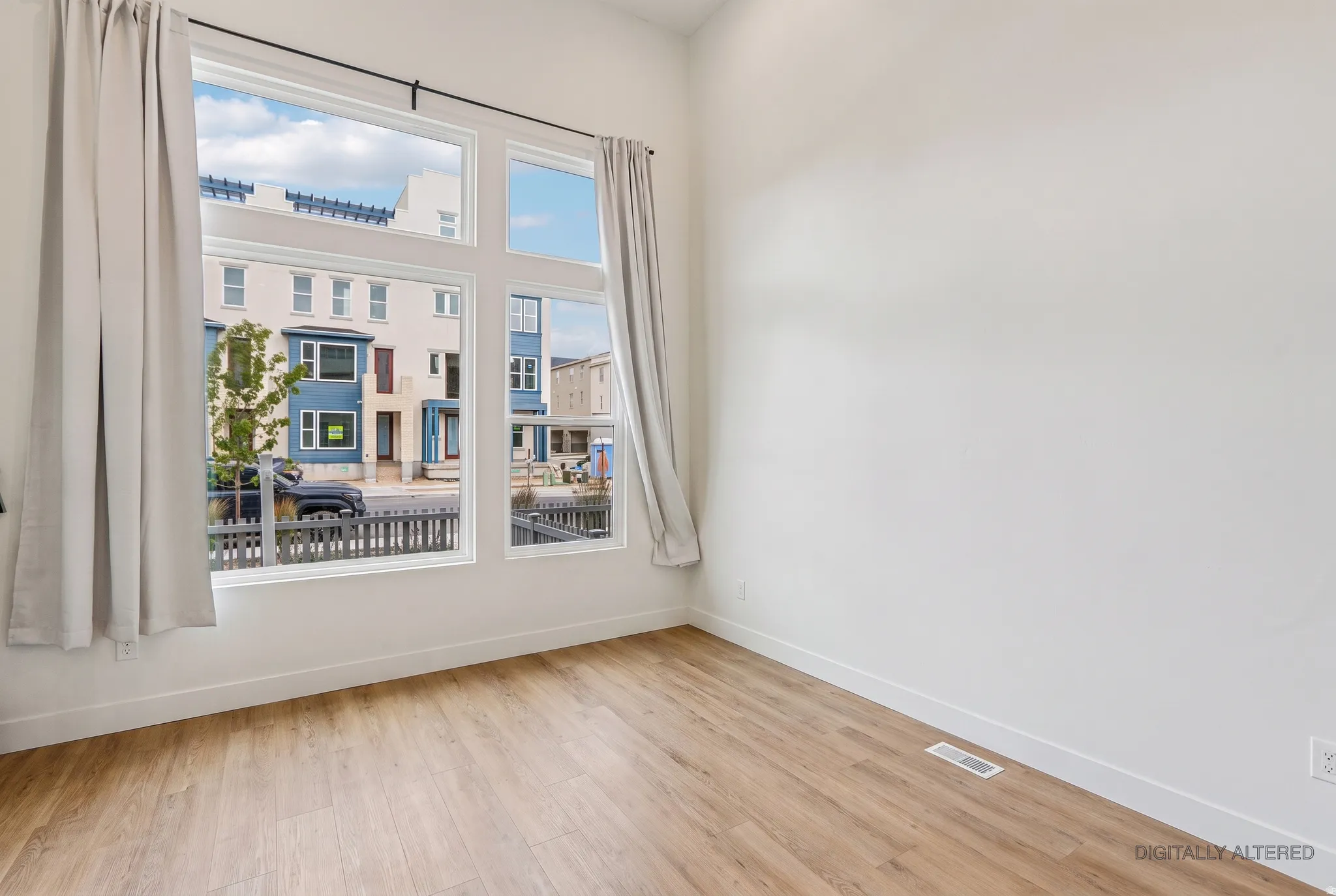 Bright corner bedroom with expansive windows framing urban streetscape, warm wood-finish flooring, and clean white walls providing a neutral backdrop for furnishings