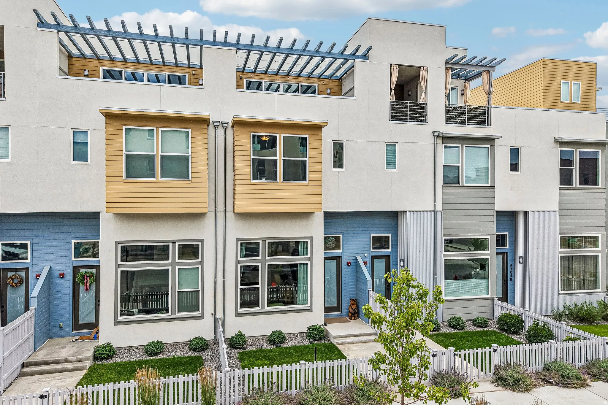 Contemporary exterior featuring multi-toned siding, expansive windows, pergolas, and a white picket fence