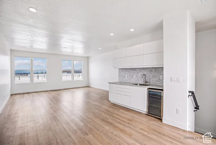 Open-concept living and kitchen space featuring gleaming wood-finish flooring throughout, expansive windows with natural light, white cabinetry with integrated appliances, gray tile backsplash, stainless steel sink and faucet, and recessed ceiling lightin