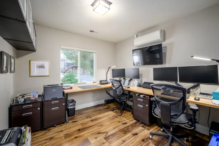 Dedicated home office featuring wood-finish flooring, dual monitor setup with built-in desk workspace, ergonomic seating, abundant natural light from large windows, and wall-mounted flat-screen display