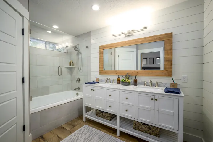 Dual vanity with a stone countertop, wood-finish flooring, a glass-enclosed shower-tub combination, and horizontal plank wall paneling