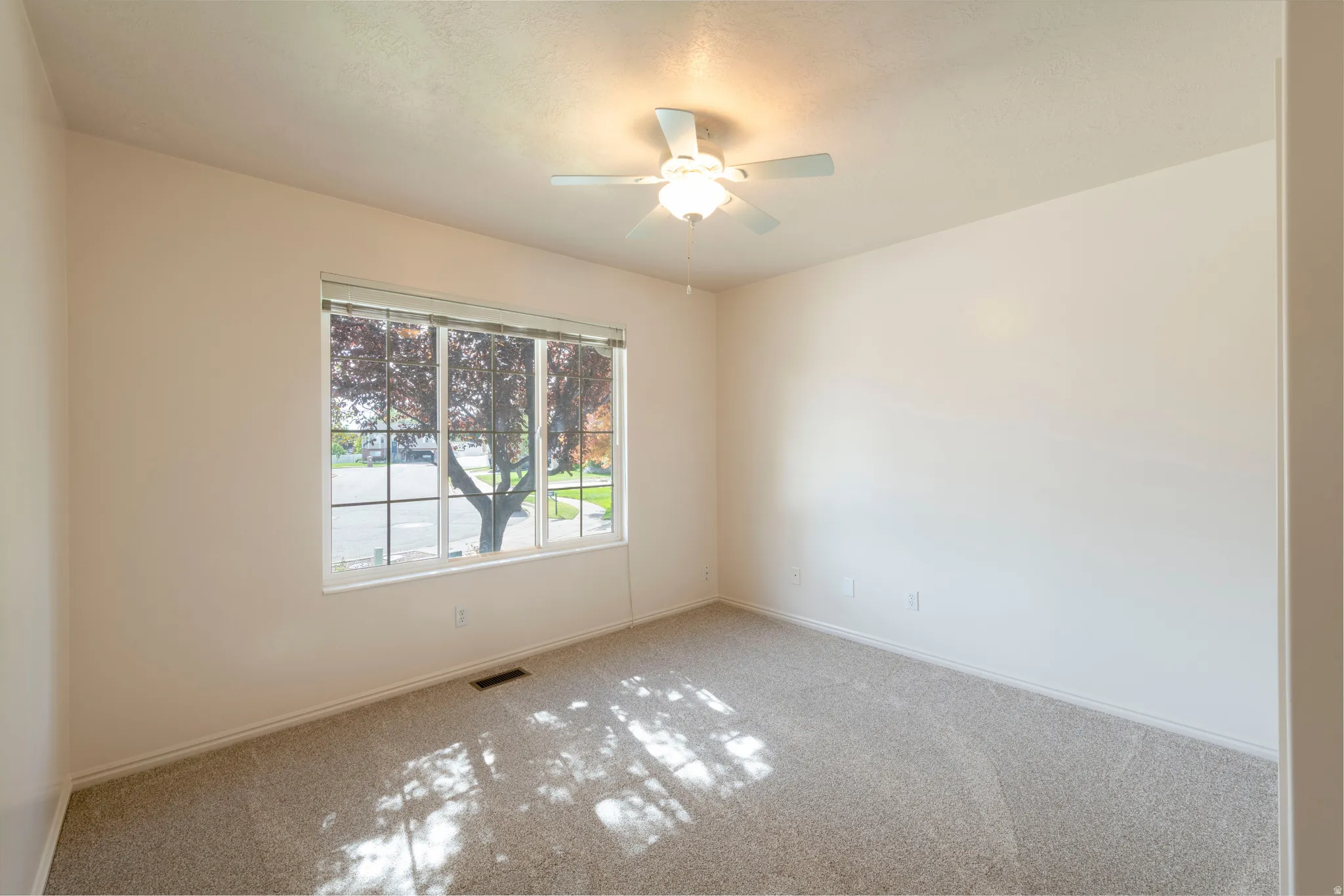 Neutral wall paint, plush carpeting, ceiling fan with integrated lighting, and a large window with horizontal blinds