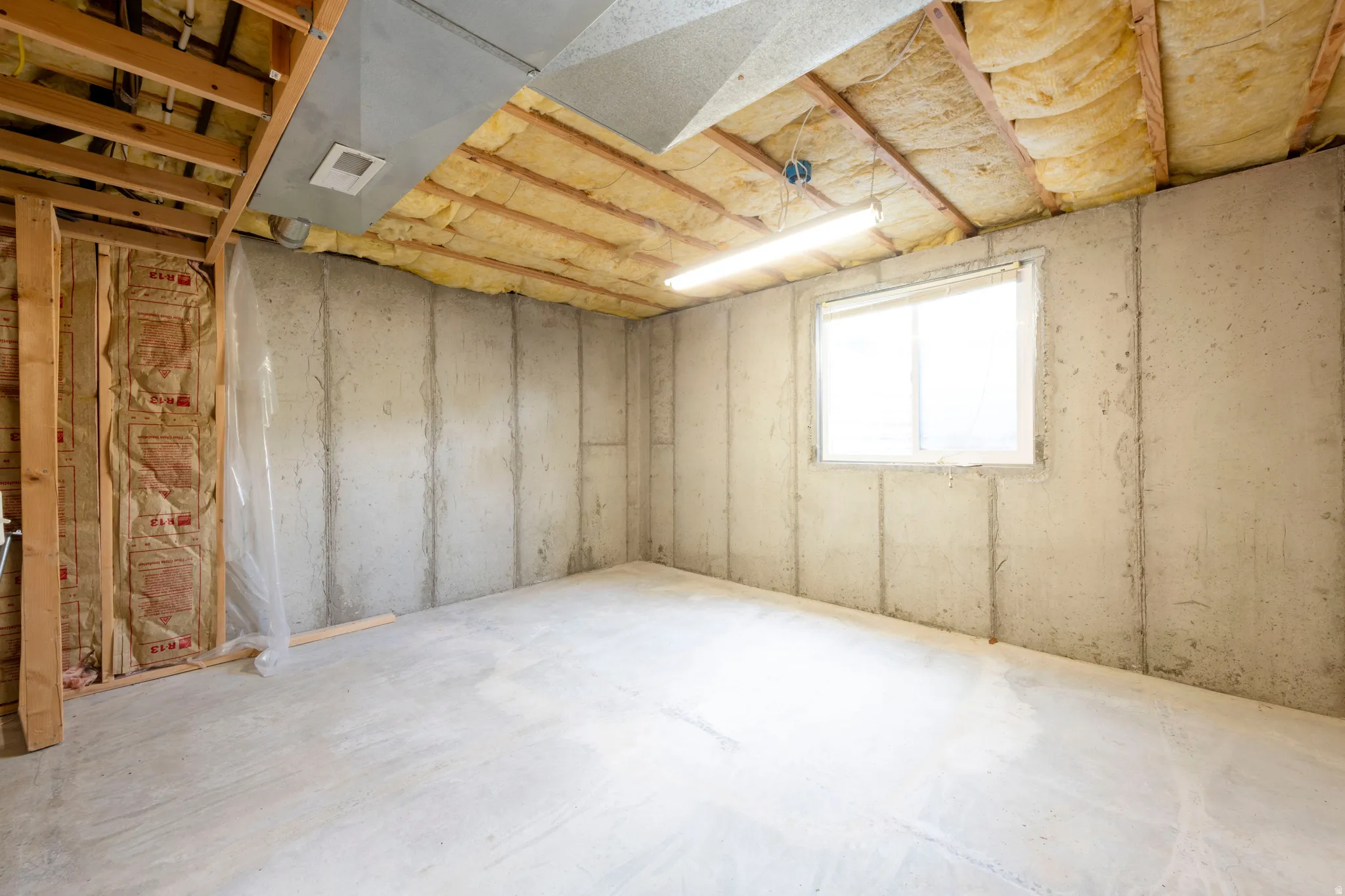 Unfinished basement featuring concrete walls and flooring, a window, and exposed ceiling joists with insulation
