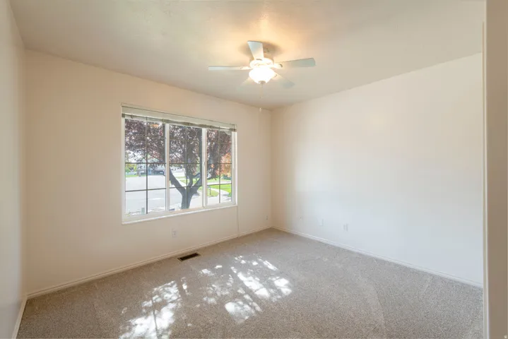 Neutral wall paint, plush carpeting, ceiling fan with integrated lighting, and a large window with horizontal blinds