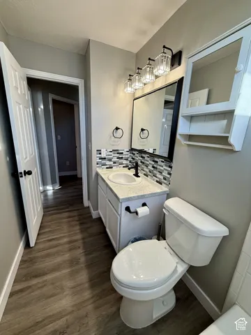 Wood-finish flooring, white vanity with a light-toned countertop, mosaic tile backsplash, and a four-light vanity fixture