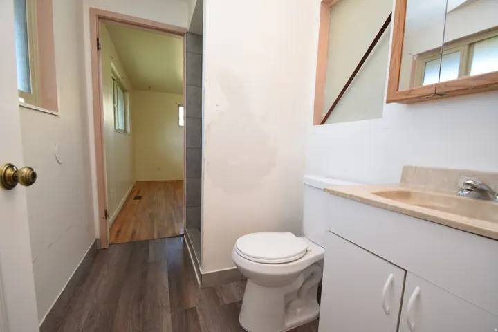 Wood-finish flooring, white vanity with integrated sink, wall-mounted mirror with wood-frame, and white toilet
