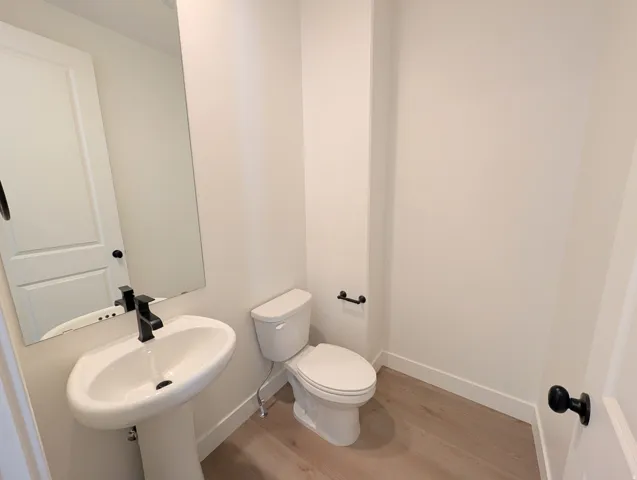 Main level half bathroom with Pedestal sink with a matte black faucet, rectangular mirror, light wood-finish flooring, and a two-piece toilet