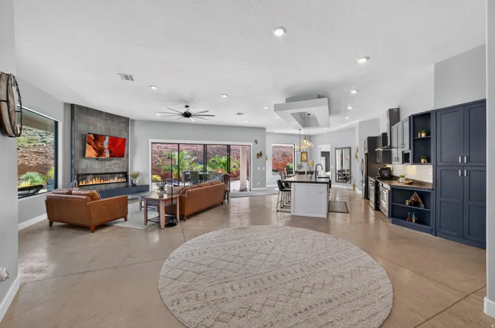 Expansive concrete flooring throughout, a modern linear fireplace with a stone surround, a contemporary ceiling fan, recessed lighting, and a kitchen featuring dark cabinetry and a center island