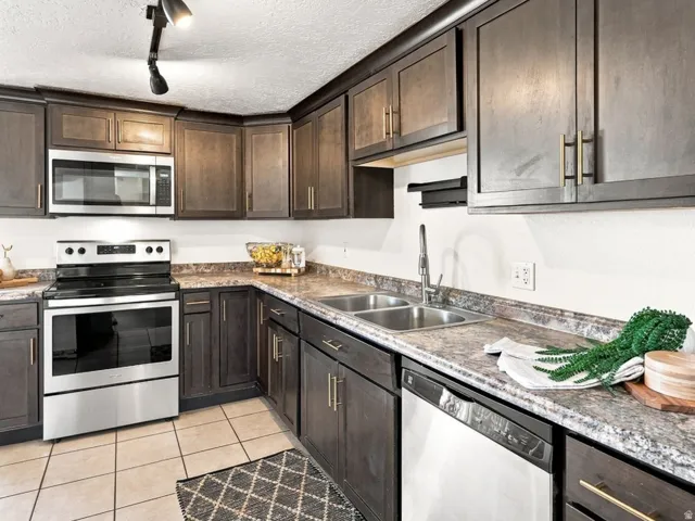 Dark wood-finish cabinetry with gold-tone hardware, stainless steel appliances, dual basin sink, light-toned tiled flooring, and speckled countertops