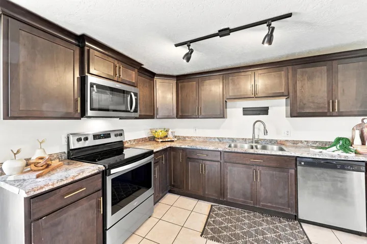 Dark wood-finish cabinetry with gold-tone hardware, a stainless steel appliance suite, light-toned patterned countertops, a stainless steel double basin sink with a gooseneck faucet, and track lighting