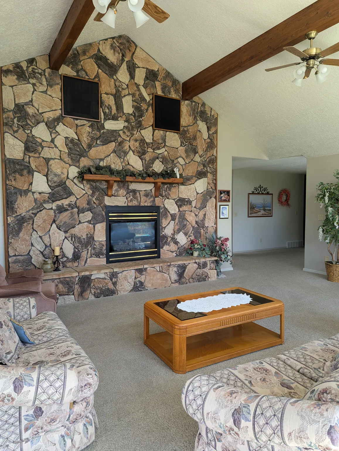 Vaulted ceiling with exposed wood beams, stone-faced fireplace with hearth, built-in fireplace insert, and ceiling fans