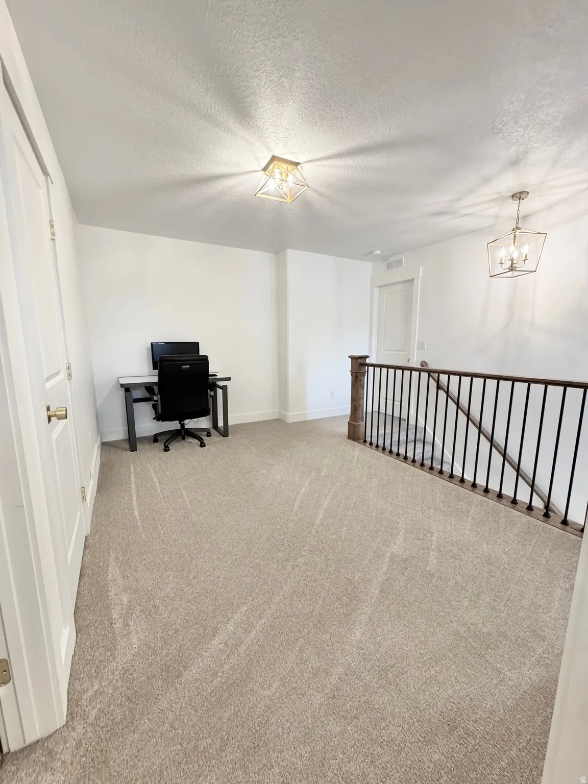 Carpeted open area with a decorative gold-tone flush mount light fixture and a wrought iron and wood banister