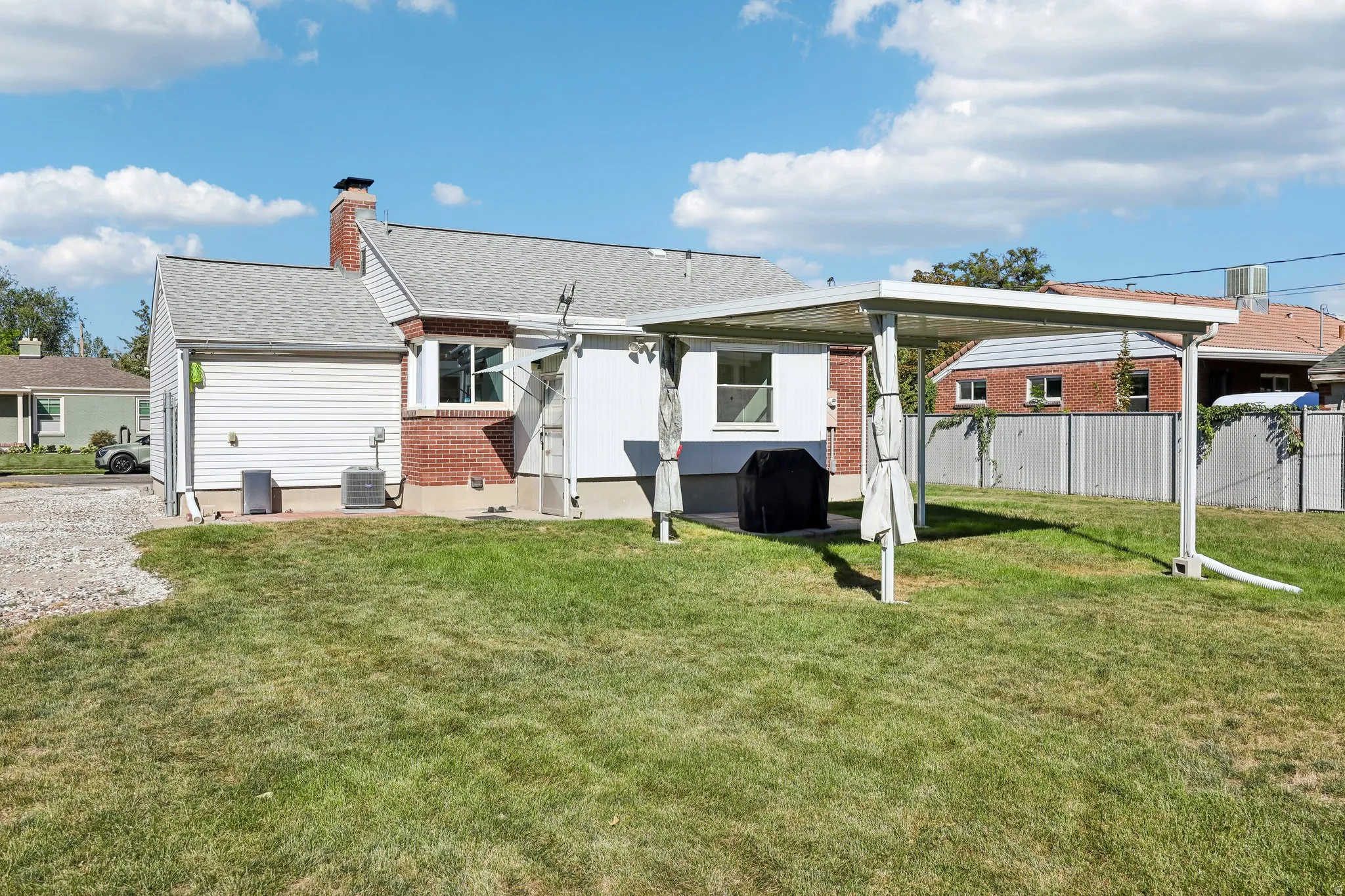 Expansive grassy yard with a covered patio area, brick and siding exterior, and a gravel side path