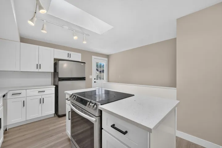 White shaker-style cabinetry with matte black hardware, light-toned countertops, stainless steel appliances, wood-finish flooring, and a skylight
