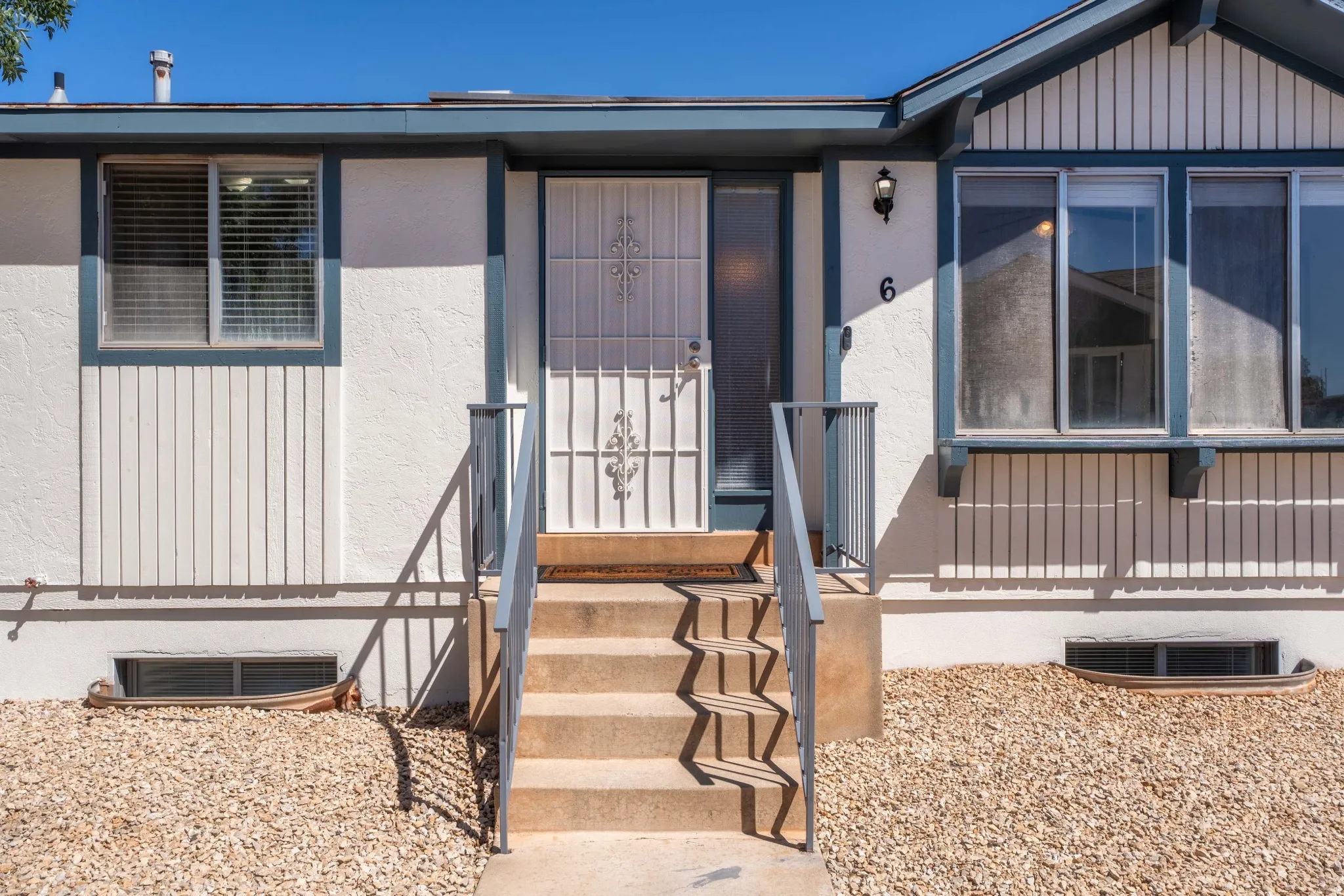 Stucco exterior with vertical paneling accents, front entry with security screen door, concrete steps with metal railings, and a gravel-covered yard