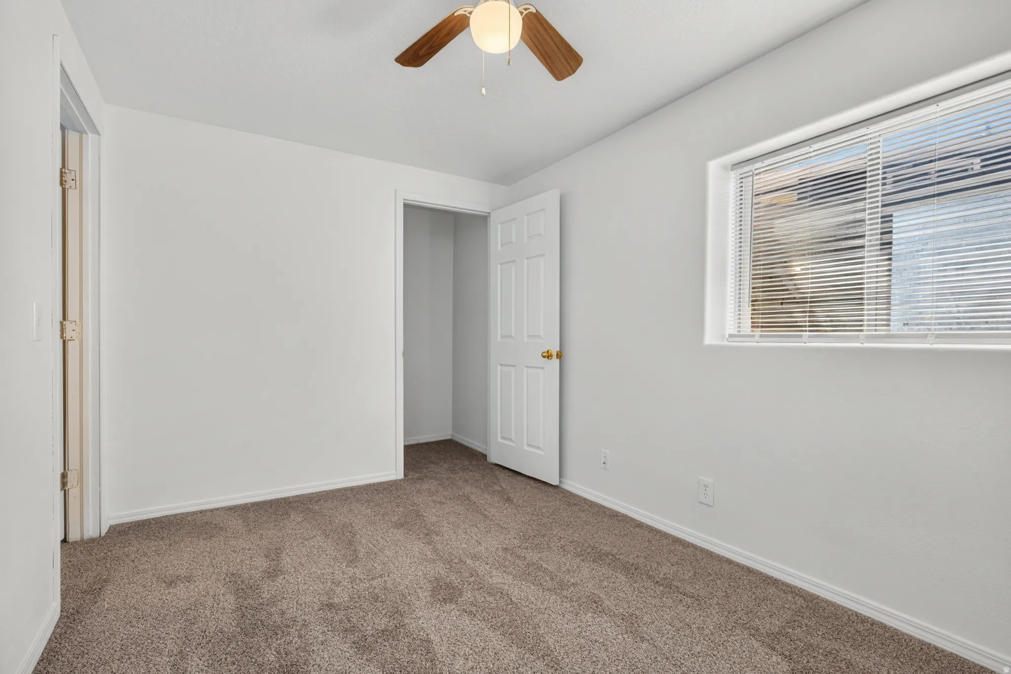Plush, neutral-toned carpet throughout, a white paneled door, a window with horizontal blinds, a ceiling fan with integrated lighting, and a recessed closet space