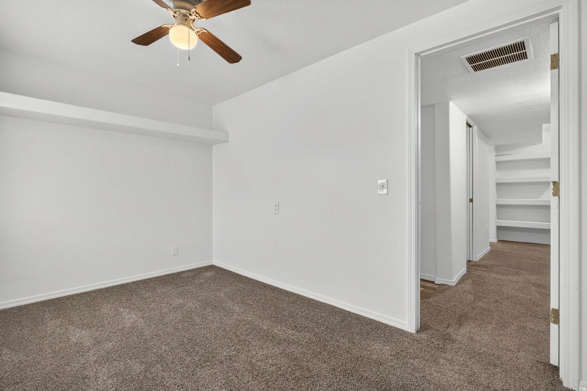 Room featuring neutral carpeting, white walls, a ceiling fan with light fixture, and a hallway with built-in shelving