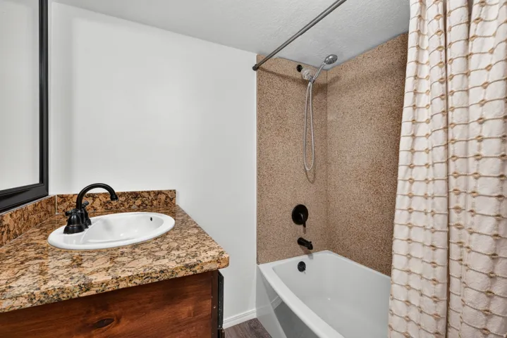 Tub and shower combination with a handheld shower head, a single basin sink, a rich wood-finish vanity, and a speckled countertop